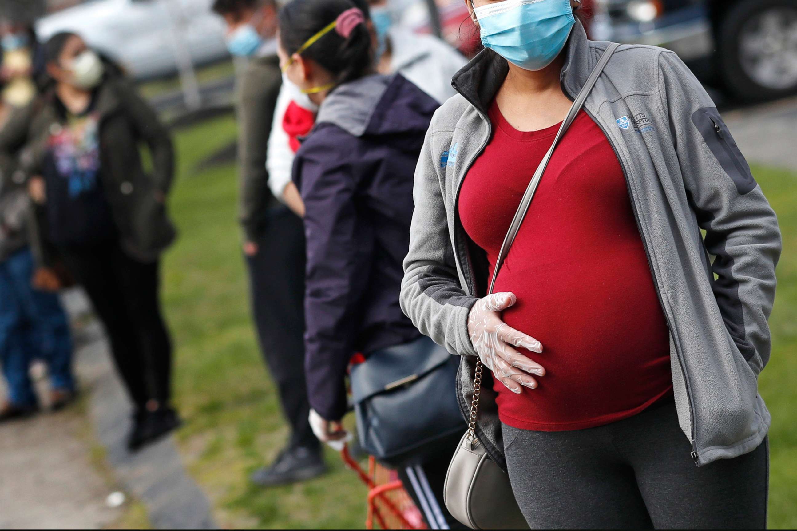 PHOTO: In this Thursday, May 7, 2020 file photo, a pregnant woman wearing a face mask and gloves holds her belly as she waits in line for groceries with hundreds during a food pantry at St. Mary's Church in Waltham, Mass.