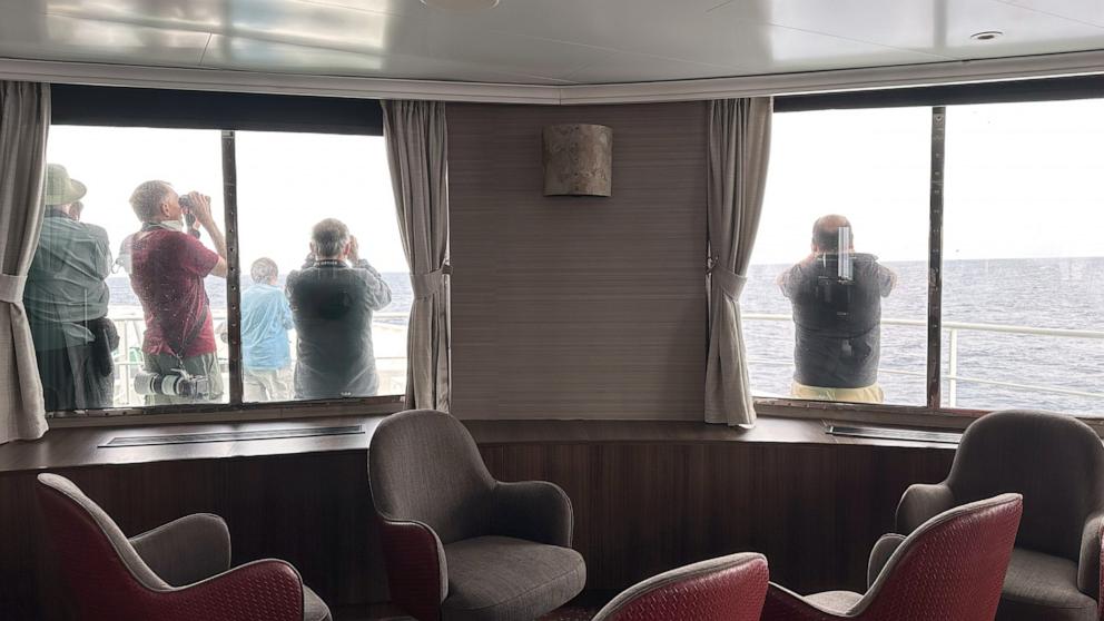 PHOTO: Passengers on the hantavirus-stricken cruise ship, MV Hondius, scan the horizon with binoculars during their voyage to Spain's port of Tenerife, May 6, 2026. 