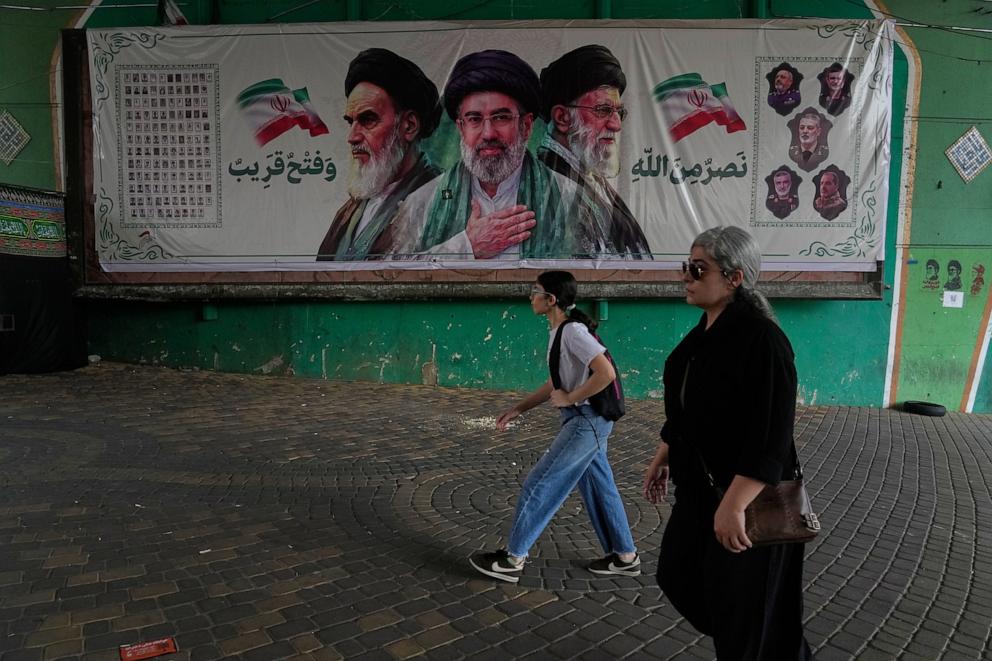 PHOTO: Women walk past a banner showing portraits of Ayatollah Ruhollah Khomeini (L), Supreme Leader Ayatollah Mojtaba Khamenei (C), and the late Supreme Leader Ayatollah Ali Khamenei, under a bridge in northern Tehran, Iran, on Aug. 2, 2026.