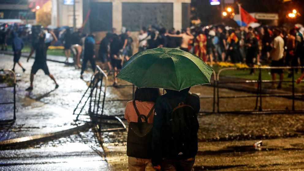 A couple under an umbrella watch as people throw stones at the police and at the parliament building in Skopje, North Macedonia, during a protest late Tuesday, July 5, 2022. Violent protests erupted in North Macedonia's capital, Skopje, where demonst