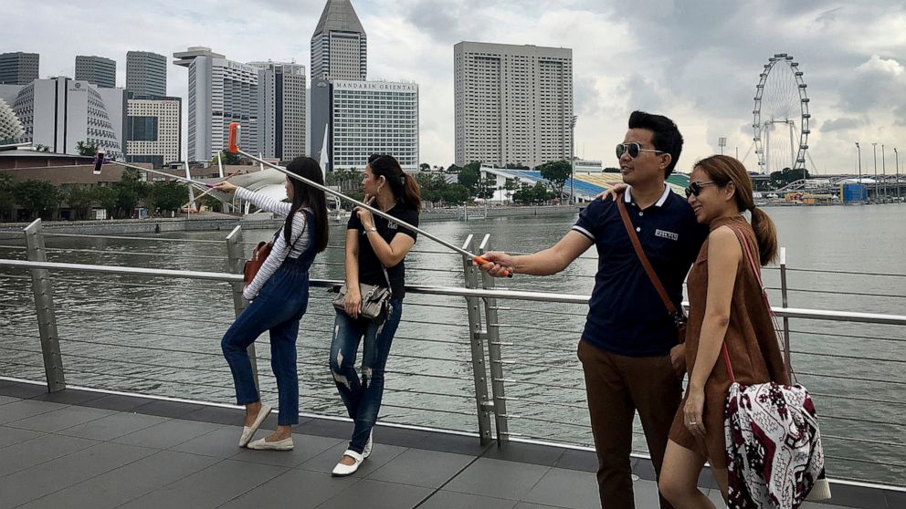FILE - In this Jan. 20, 2016, file photo, tourists use selfie sticks to take photos of themselves with their smart phones at one of Singapore's popular tourists spots with the financial skyline and hotels in the background in Singapore. The World Eco