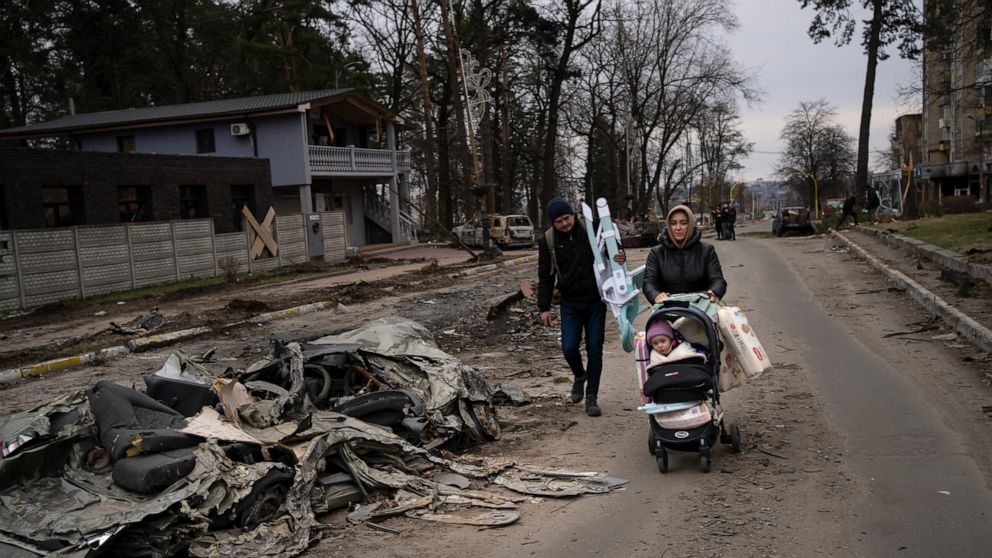 A family walks pass a car crushed by a Russian tank in Bucha, in the outskirts of Kyiv, Ukraine, Tuesday, April 5, 2022. Ukraine's president planned to address the U.N.'s most powerful body on Tuesday after even more grisly evidence emerged of civili