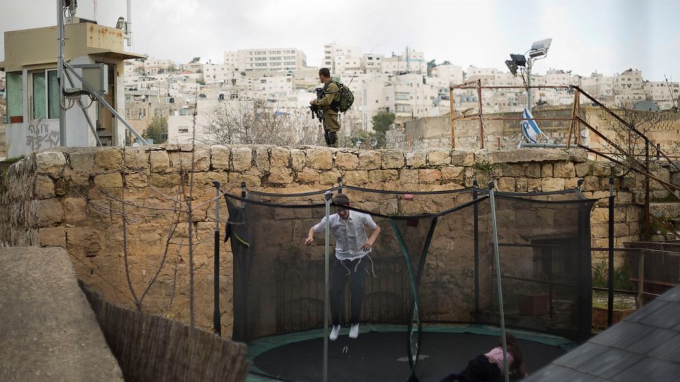 FILE - In this March 7, 2019, file photo, settlers jump on a trampoline as an Israeli solider stands guard in the Israeli controlled part of the West Bank city of Hebron. Israel's premier human rights group has begun describing both Israel and its co