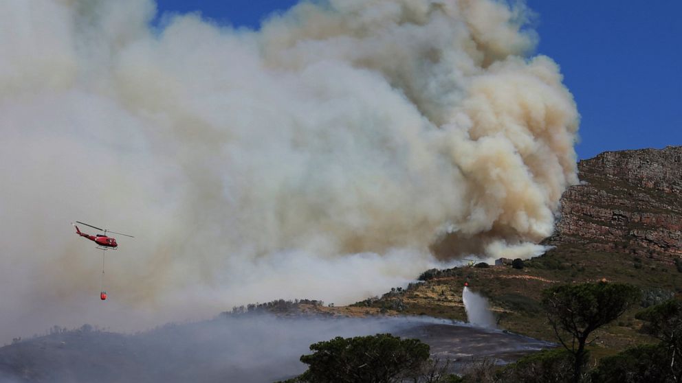 A helicopter drops water over a fire at Rhodes Memorial on Table Mountain, Cape Town, South Africa, Sunday, April 18, 2021. A wildfire raging on the slopes of the mountain forced the evacuation of students from the University of Cape Town. (AP Photo/