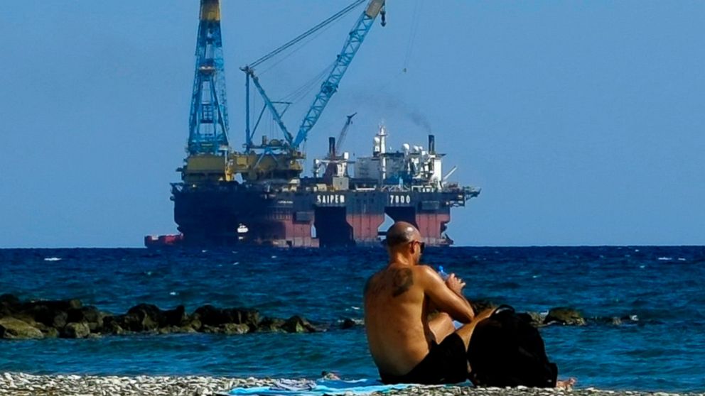 FILE - In this Sunday, Oct. 15, 2017 file photo, a man sits on a beach as a drilling platform is seen in the background outside from Larnaca port, in the eastern Mediterranean island of Cyprus. Cyprus' energy minister says ExxonMobil has discovered t