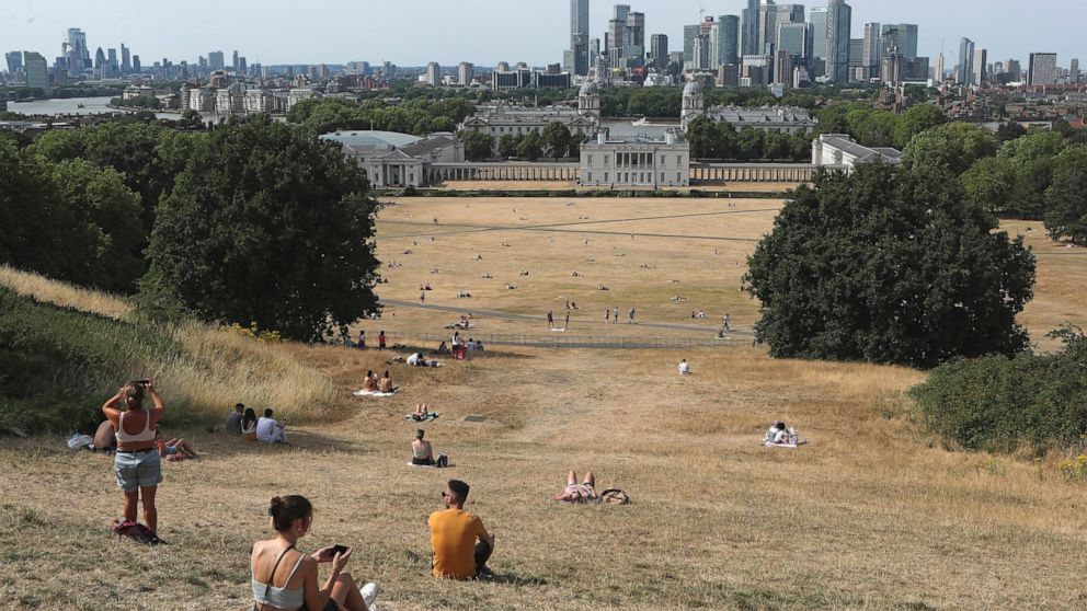 People sit on the sun-parched grass in Greenwich Park with the Maritime museum and Canary Wharf financial district in the background in London, Sunday July 17, 2022. Britain's weather agency has issued its first-ever "red warning" of extreme heat for