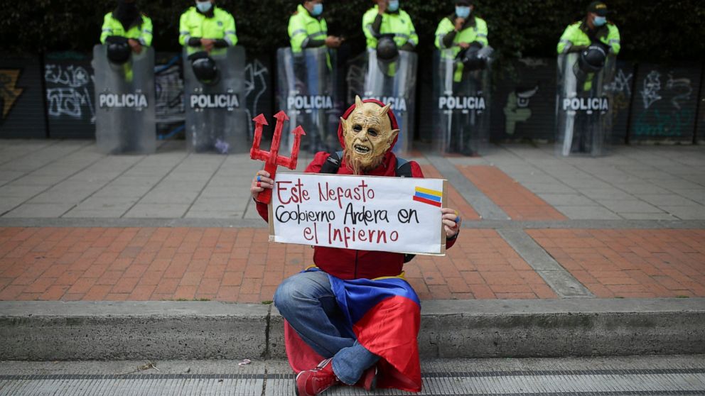 A man in a devil costume holds the Spanish sign "This disastrous government will burn in hell" in front of police standing on the sidelines of an anti-government march in Bogota, Colombia, Wednesday, May 19, 2021. Colombians have taken to the streets