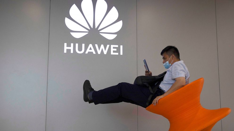 FILE - A man wearing a mask to curb the spread of the coronavirus sits near a Huawei store logo in Beijing on Friday, July 31, 2020. Chinese telecoms equipment and smartphone maker Huawei said Thursday its sales fell 14% in the last quarter from a ye