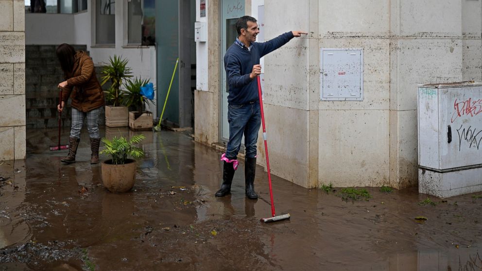 A man sweeping water from a building shows passers by the height that the water reached when the street was flooded overnight in Alges, just outside Lisbon, Tuesday, Dec. 13, 2022. An Atlantic storm slammed into the Iberian peninsula, leaving behind 