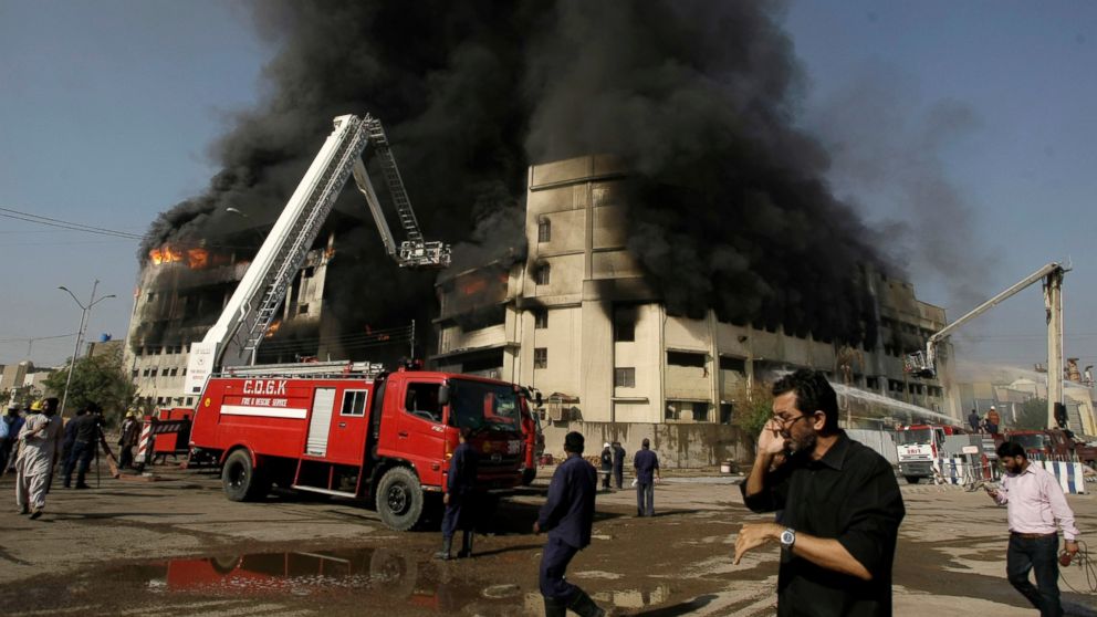 FILE - In this Sunday, Oct. 28, 2012 file photo, a man speaks on his mobile phone, as firemen extinguish a fire that erupted in a factory in Karachi, Pakistan. A German court won’t take up a civil case against a discount textile company whose clothes