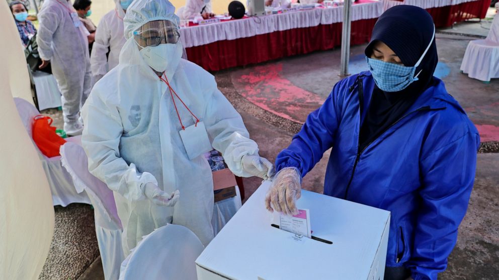 An electoral worker wearing a protective suit as a precaution against the coronavirus outbreak assists a woman to cast her ballot during the regional election at a polling station in Tangerang, Indonesia, Wednesday, Dec. 9, 2020. Indonesia pushed for