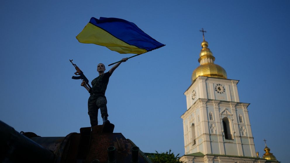 A woman brandishes the Ukrainian flag on top of a destroyed Russian tank in Kyiv, Ukraine, Friday, June 10, 2022. With war raging on fronts to the east and south, the summer of 2022 is proving bitter for the Ukrainian capital, Kyiv. The sun shines bu
