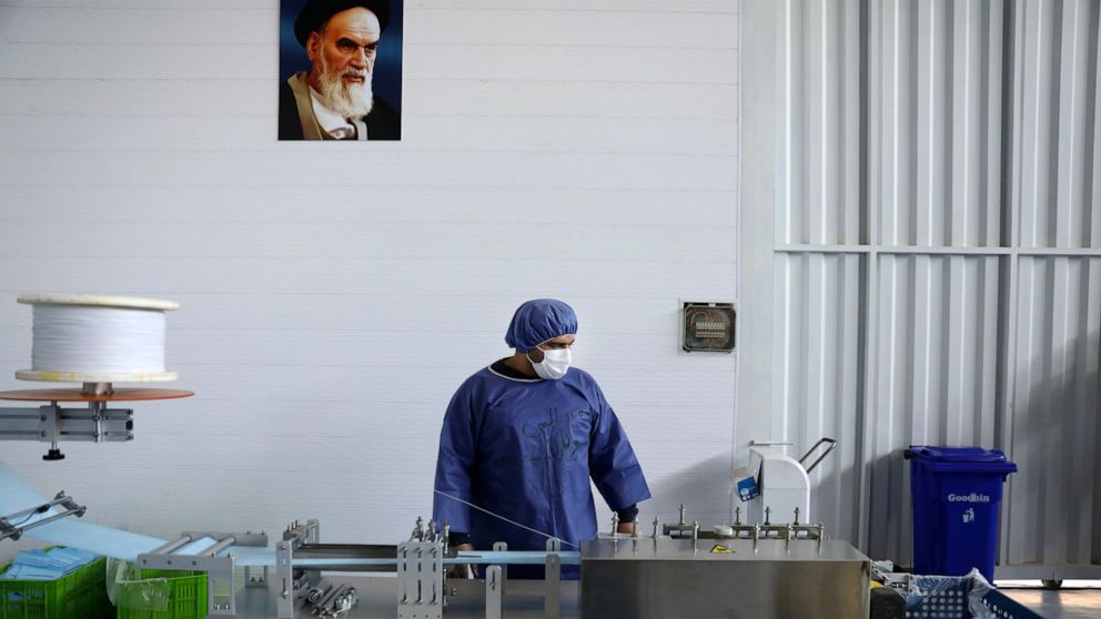 Under a portrait of the late Iranian revolutionary founder Ayatollah Khomeini, a worker wearing a protective suit controls a machine which makes face masks at a factory in Eshtehard, some 70 miles (114 kilometers) west of the capital Tehran, Iran, Tu