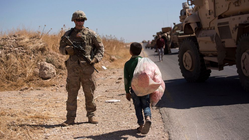 A Syrian boy selling snacks looks at a U.S. soldier standing guard during the first joint ground patrol by American and Turkish forces in the so-called "safe zone" on the Syrian side of the border with Turkey, near Tal Abyad, Syria, Sunday, Sept. 8, 