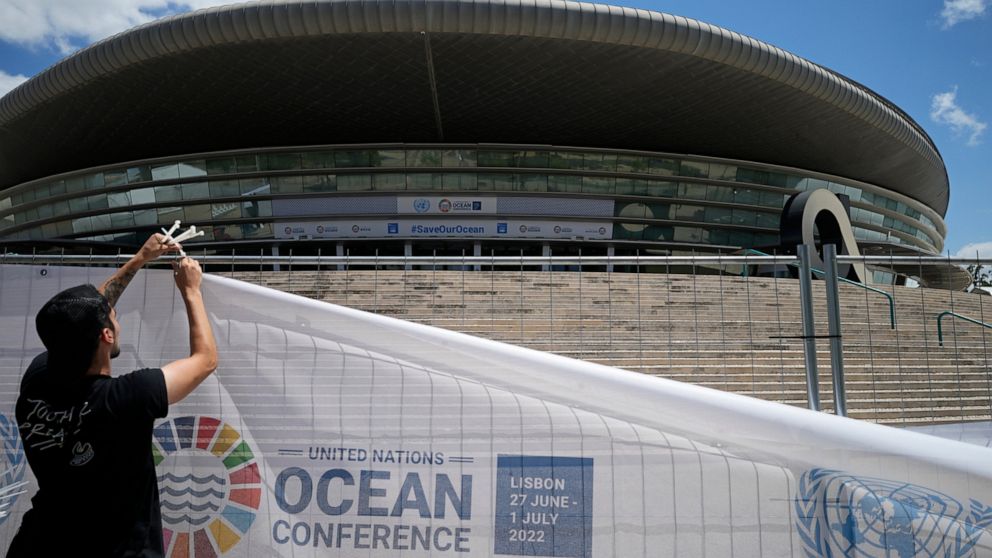 A worker hangs a banner on the fence surrounding the sports arena in Lisbon that will host the United Nations Oceans Conference, Saturday, June 25, 2022. From June 27 to July 1, the United Nations is holding its Oceans Conference in Lisbon expecting 