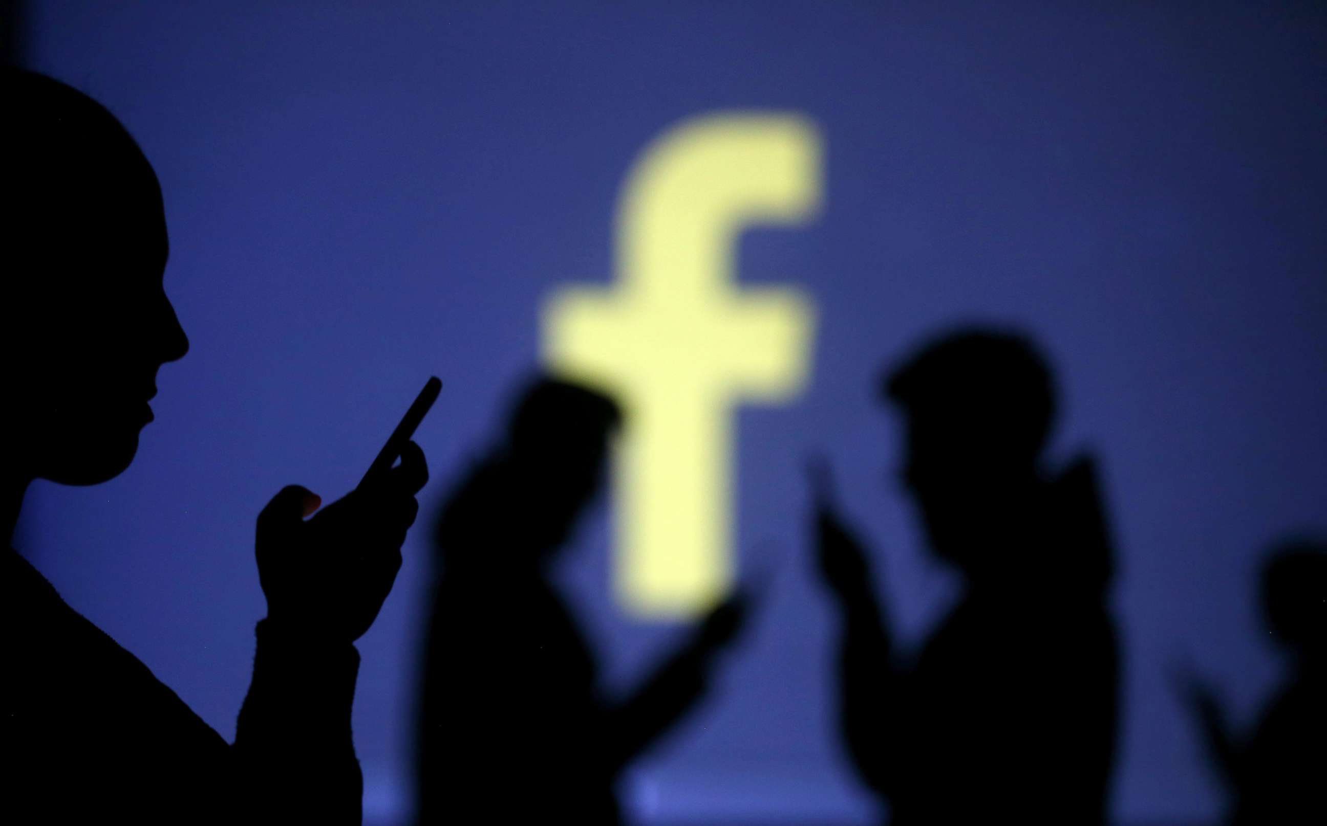PHOTO: Silhouettes of mobile users are seen next to a screen projection of Facebook logo in this picture illustration taken March 28, 2018.