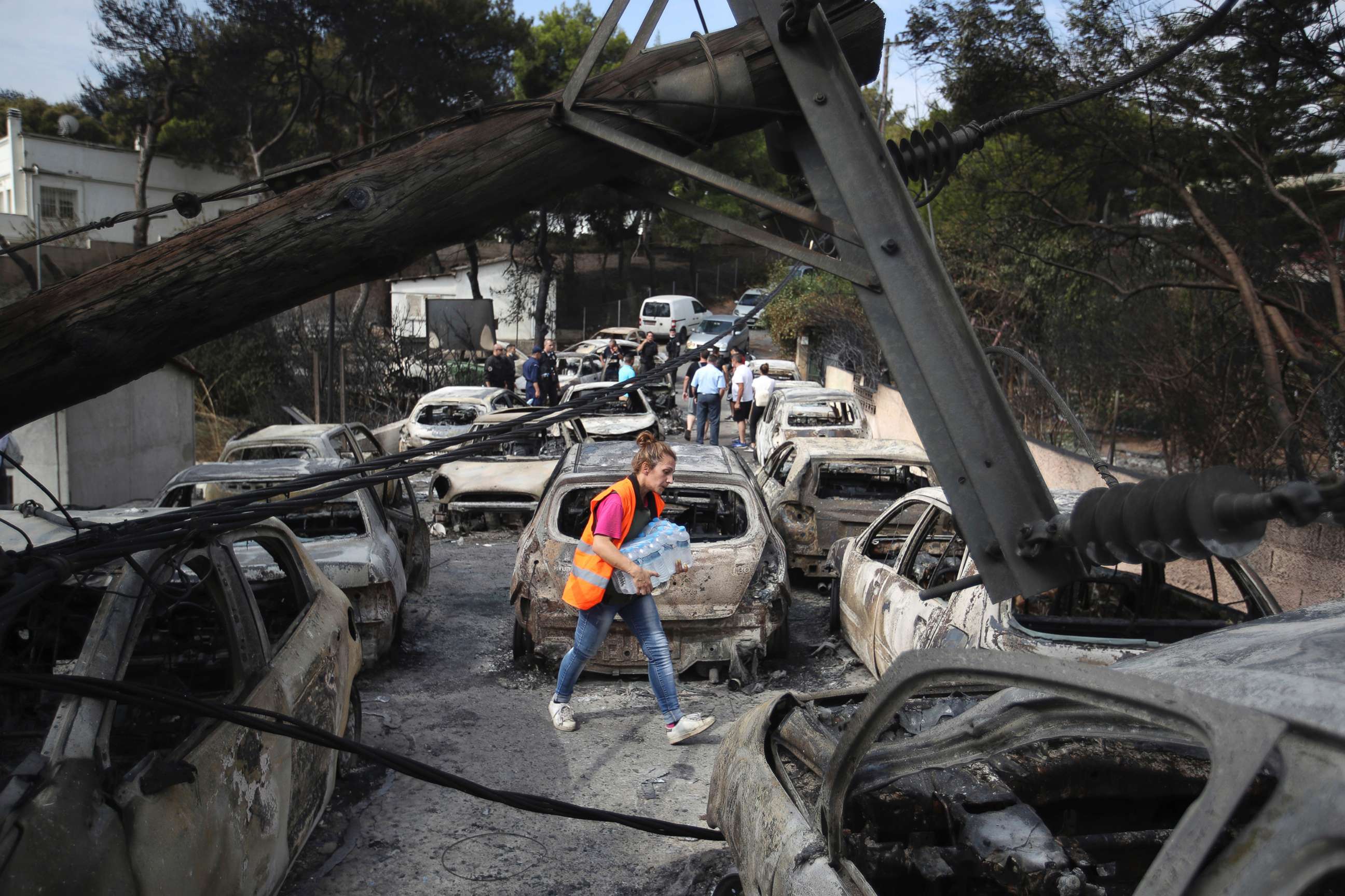 PHOTO: A woman carries bottles of water as people stand amid the charred remains of burned-out cars in Mati east of Athens, July 24, 2018.