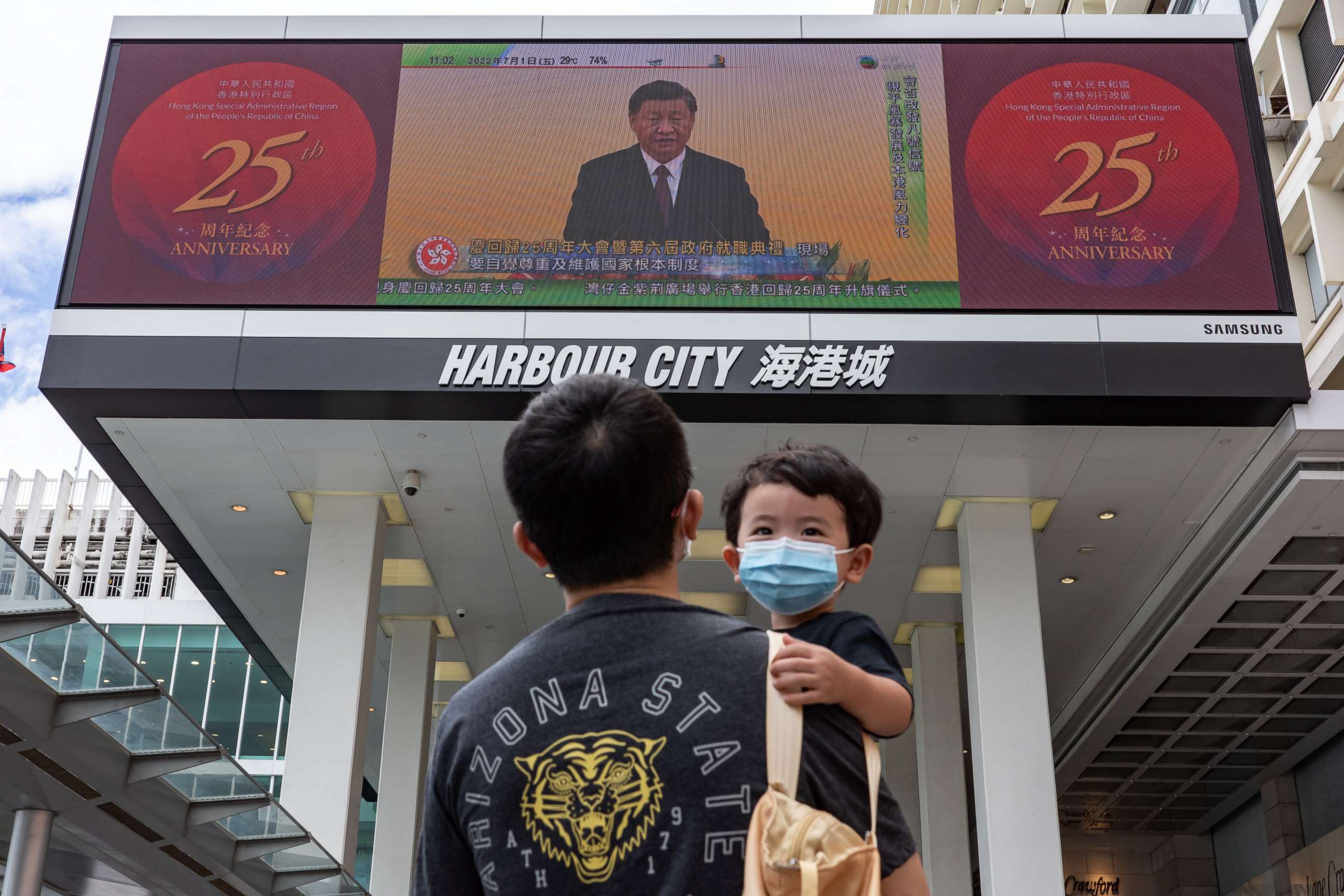 PHOTO: HONG KONG, CHINA: A man holding a child walks in front of a screen showing a live broadcast of Chinese President Xi Jinping speaking during a swearing-in ceremony for Hong Kong's new chief executive John Lee, on July 01, 2022 in Hong Kong, China. 