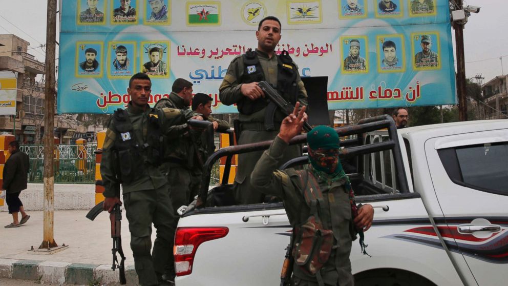 Members of the Kurdish internal security forces stand on their vehicle in front of a giant poster showing portraits of fighters killed fighting against the Islamic State group, in Manbij, Syria, March 28, 2018.