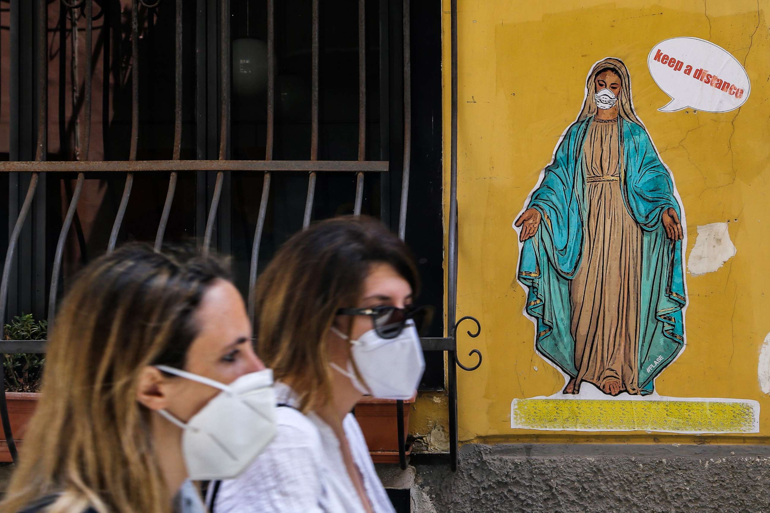 PHOTO: Women wearing a face masks walk past a mural by Art Flase depicting the Virgin Mary wearing a mask and saying "Keep a distance" in downtown Naples, Italy, on May 13, 2020.