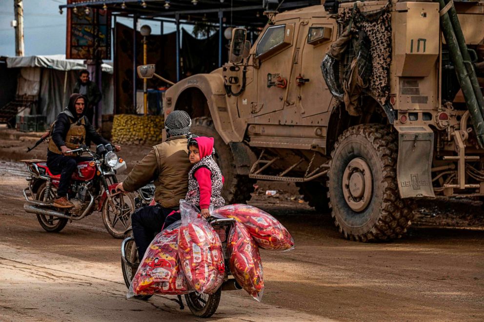 PHOTO: A man and a child ride a motorcycle loaded with bags of snacks as a patrol of U.S. military vehicles is seen in the town of Tal Tamr in the northeastern Syrian Hasakeh province along the border with Turkey on Feb. 8, 2020.