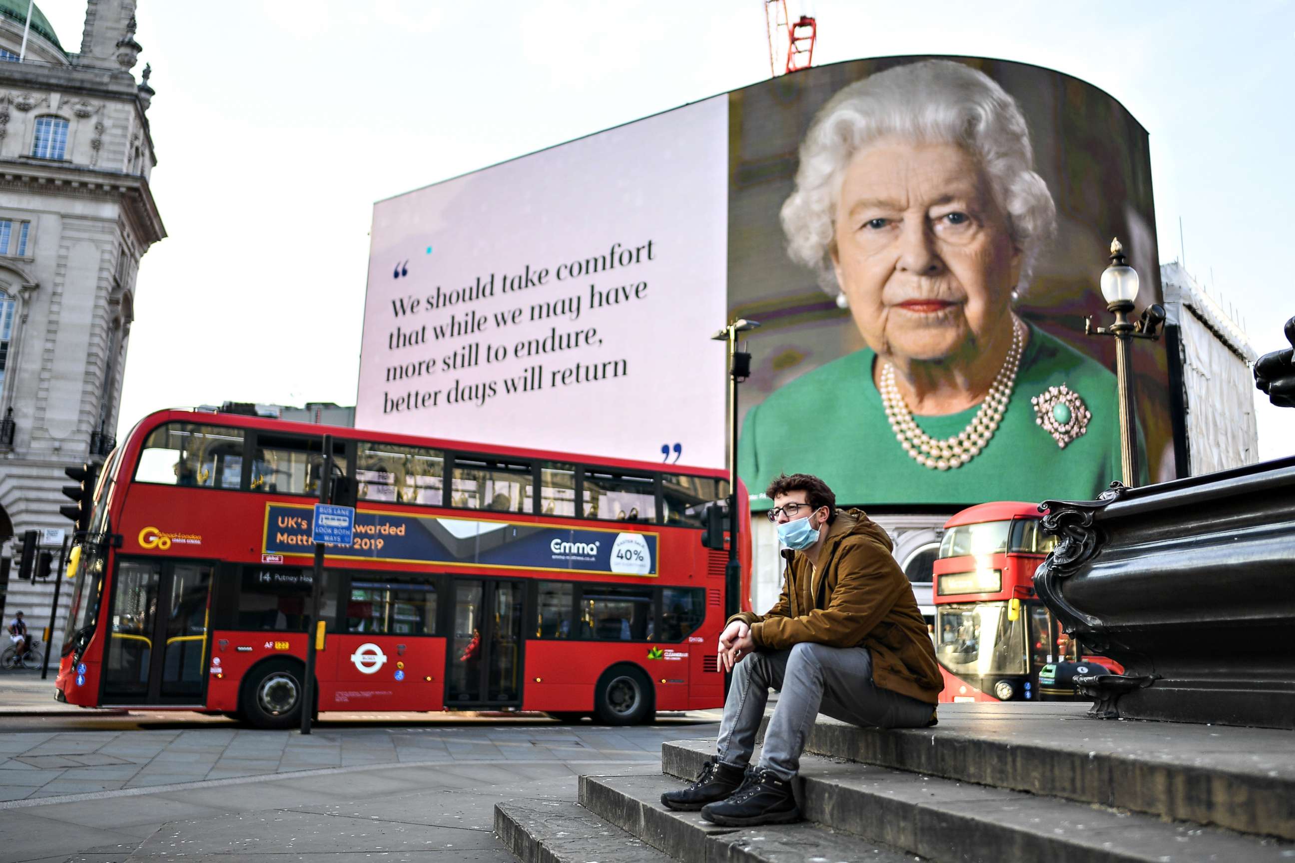 PHOTO: A man wearing a face mask sits in front of an image of Queen Elizabeth II with quotes from her broadcast to the nation on the coronavirus epidemic displayed on screens in Piccadilly Circus, on April 09, 2020, in London, England.