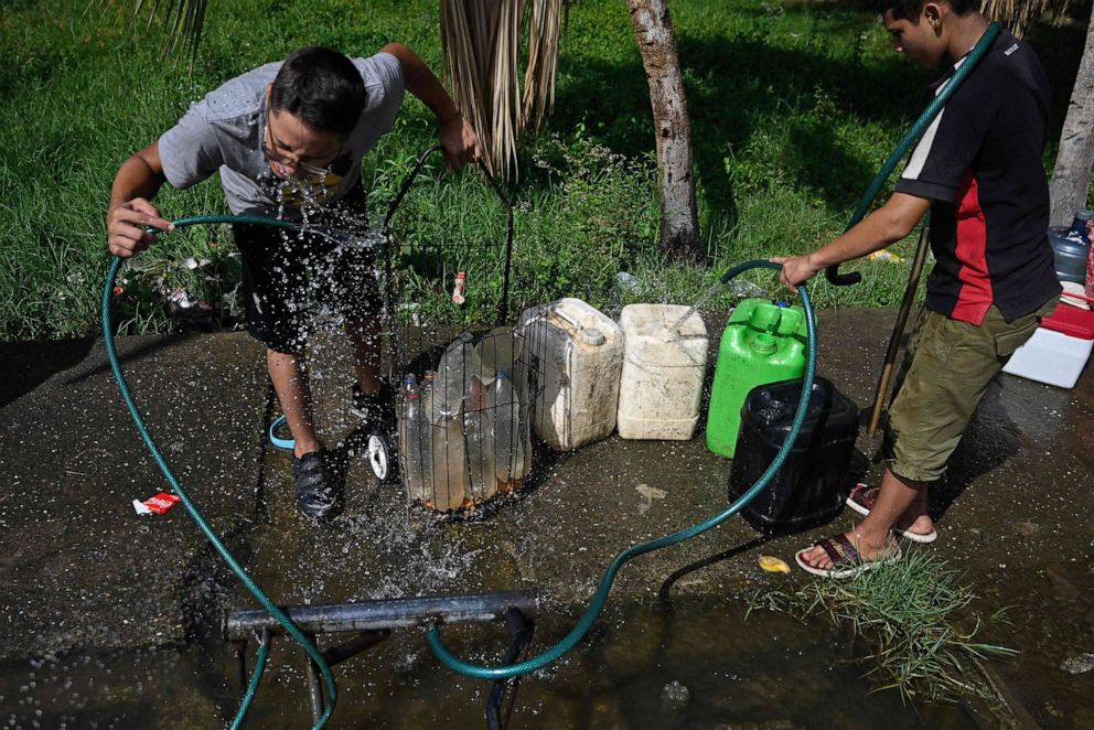 PHOTO: A boy drinks water as another fills jerry cans from a makeshift intake set on a sidewalk for people to collect water in Maracaibo, Zulia State, Venezuela, on July 23, 2019.