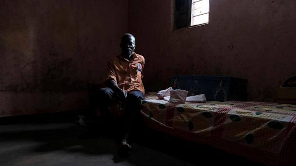 Simon Tigawalana sits on a bed as he rests his feet on a clay-based earthen floor made by EarthEnable at home on May 16, 2025, in Jinja, Uganda. (AP Photo/Hajarah Nalwadda)