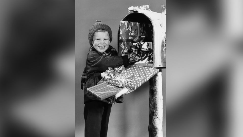 Studio portrait of a grinning young boy wearing winter clothes finding wrapped Christmas gifts in a large mailbox, circa 1955.