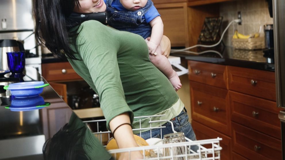 PHOTO: A busy mother holds her child while putting dishes in a dishwasher in this stock image.