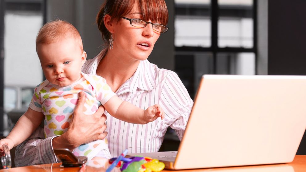 A mother holds her baby while working on a laptop in this stock photo.