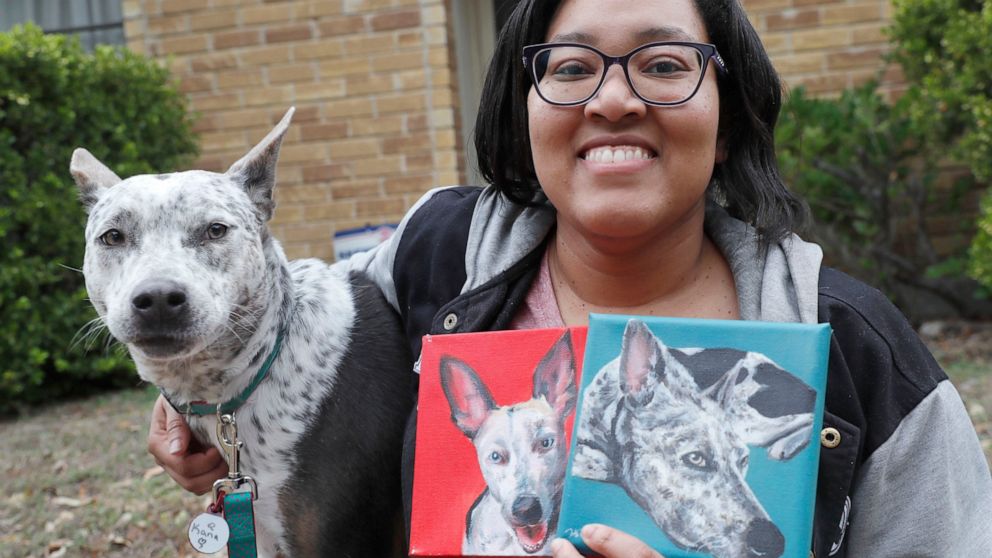 In this photo made Monday, March 2, 2020, Danielle Moore poses for photos with Kana and also paintings of the pet Australian cattle dog in Dallas. In the dog-eat-dog world of online shopping, Chewy has an unusual plan to fend off Amazon: turning pets