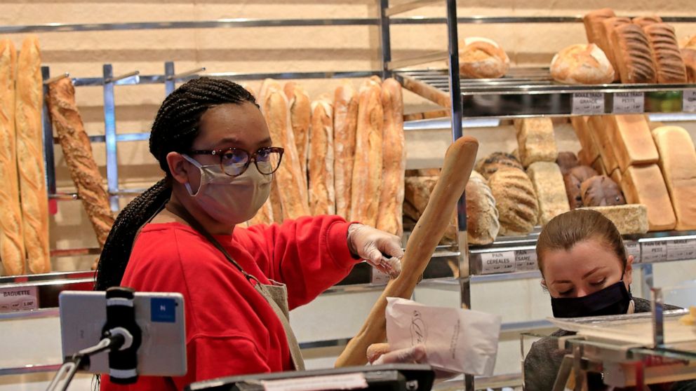 A vendor wearing a protective mask puts a baguette in a paper bag at a bakery in Paris, Monday, March 23, 2020. For most people, the new coronavirus causes only mild or moderate symptoms, such as fever and cough. For some, especially older adults and
