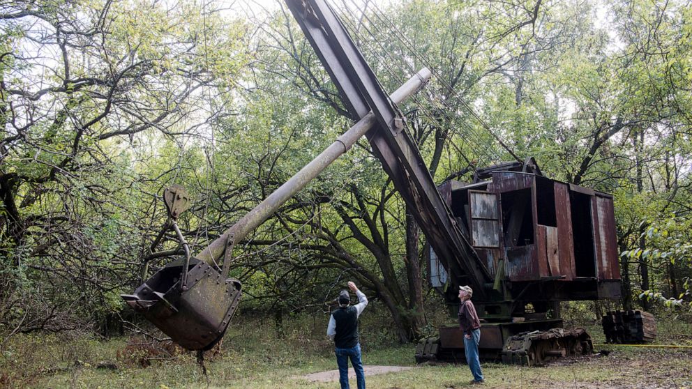 FILE--In this Oct. 16, 2018 file photo, Jim Lovell, left, and Carmen Boccia look over the Markley shovel where it has been for 70 years in nearby Cherokee, Kan. The historic coal mining shovel that was hidden under bramble for more than 70 years has 