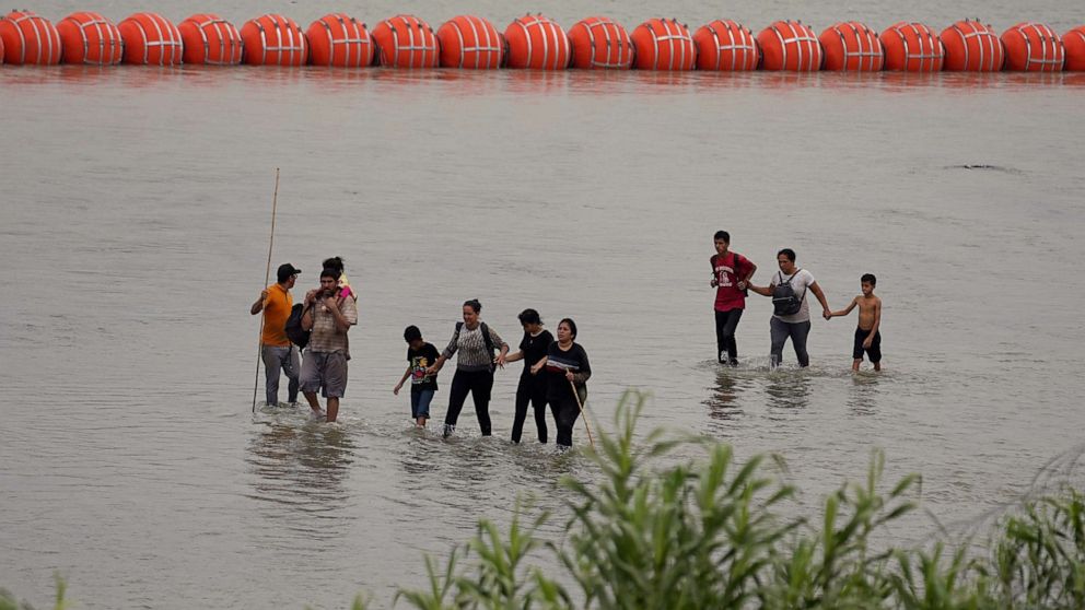 PHOTO: Migrants who crossed the Rio Grande from Mexico walk past large buoys being deployed as a border barrier on the river in Eagle Pass, Texas, July 12, 2023.