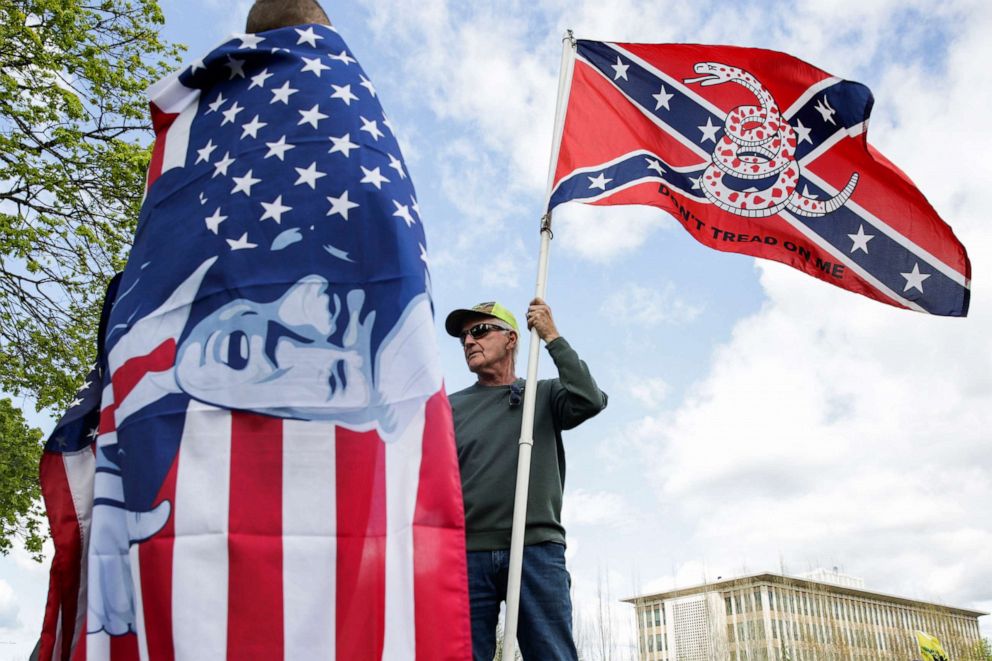 PHOTO: Keith Weber holds a flag that combines a Gadsden flag from the American Revolution with a Confederate flag from the American Civil War as he talks to protesters holding flags with President Donald Trump on them  in Olympia, Wash., April 19, 2020. 