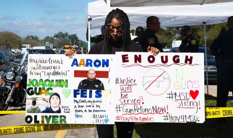 PHOTO: Tyra Hemats, 19, a 12th grader at Marjory Stoneman Douglas High School, stands with signs, one advocating for gun reform and another honoring her coach and friend, both of whom were killed in Wednesday's shooting, on Feb. 15, 2018. 