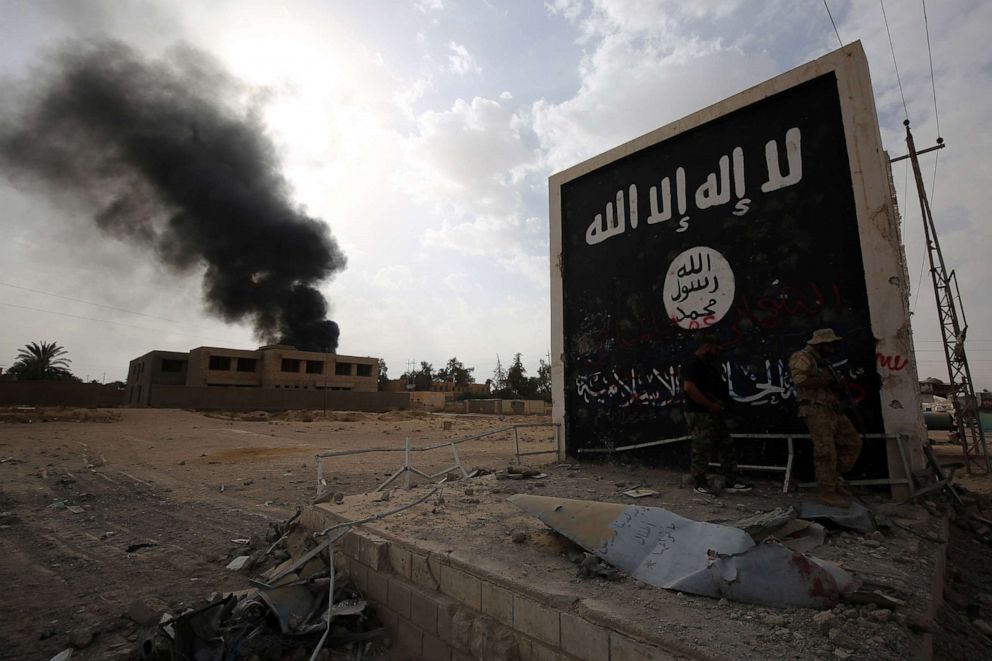 PHOTO: In this Nov. 3, 3017, file photo, Iraqi fighters of the Hashed al-Shaabi stand next to a wall bearing the Islamic State (IS) group flag as they enter the city of al-Qaim, in Iraq's western Anbar province near the Syrian border.