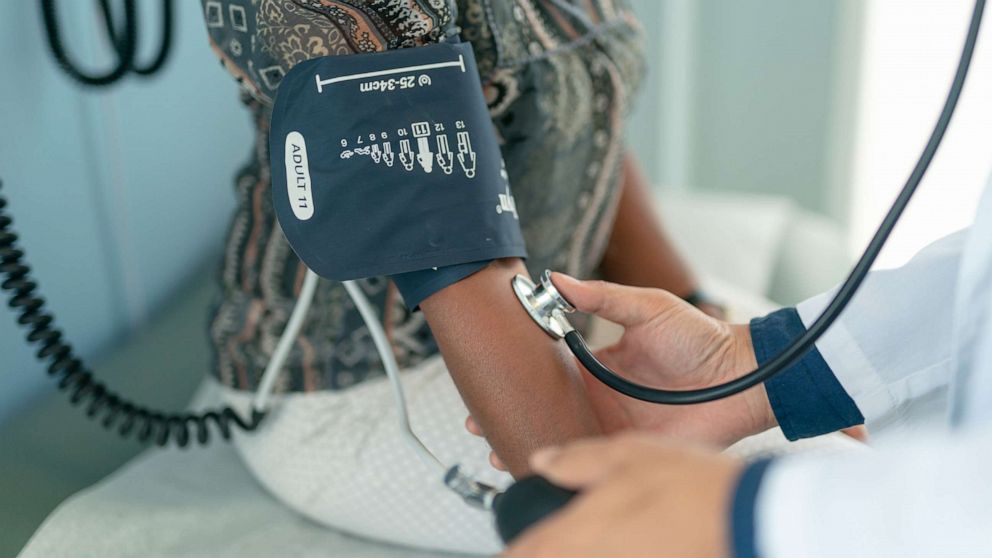 PHOTO: A woman sits on an examination table in a clinic while a health care professional checks her blood pressure.