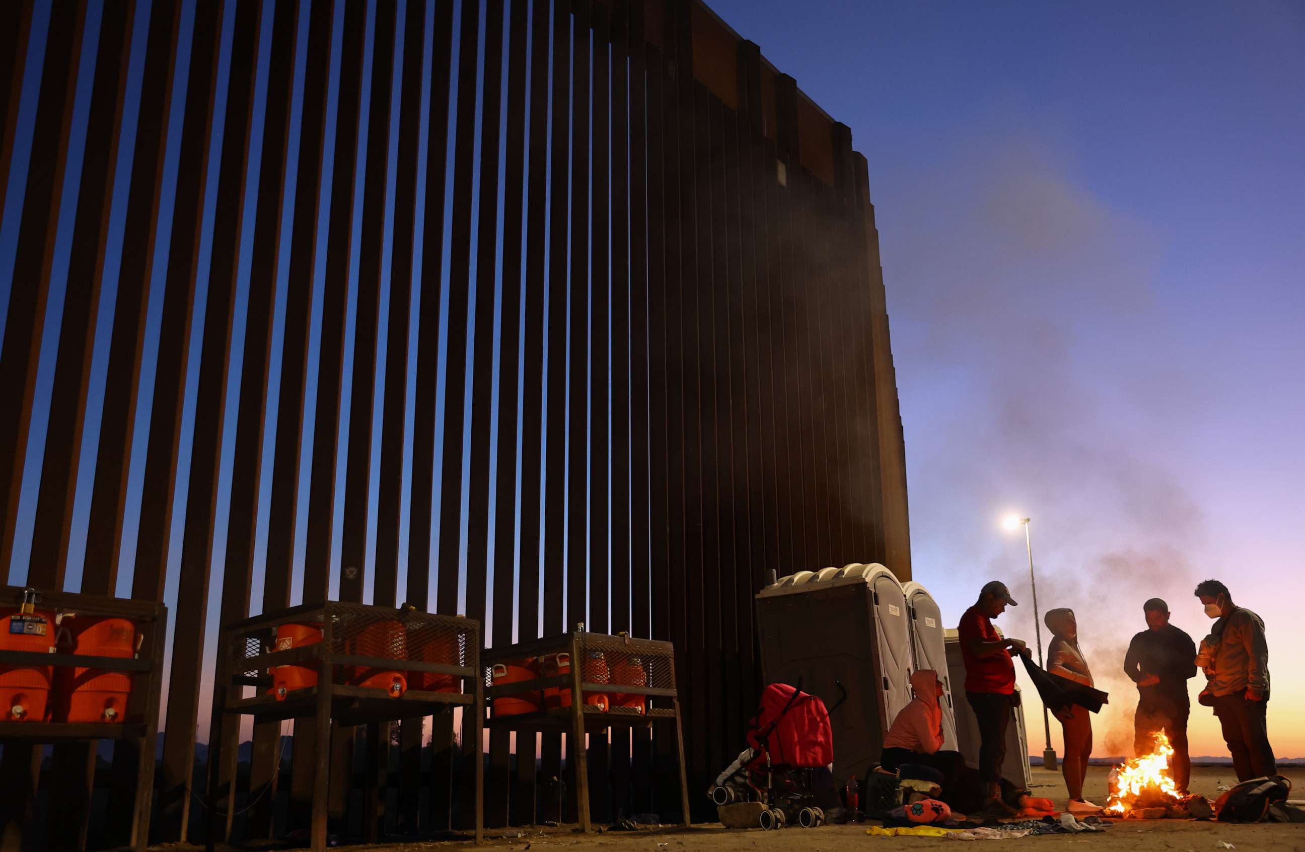 PHOTO: Immigrants from Cuba and Venezuela warm themselves by a fire before sunrise along the U.S.-Mexico border barrier as they await processing by the U.S. Border Patrol after crossing from Mexico on May 22, 2022 in Yuma, Ariz.