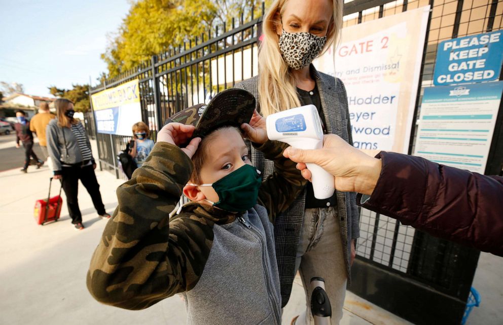 PHOTO: First grade student Baron White gets his hat into position to have his temperature taken by teachers aide Firoozeh Borjian as he arrives with his Mom Alison White for class in Redondo Beach, Calif., Feb. 2, 2021.