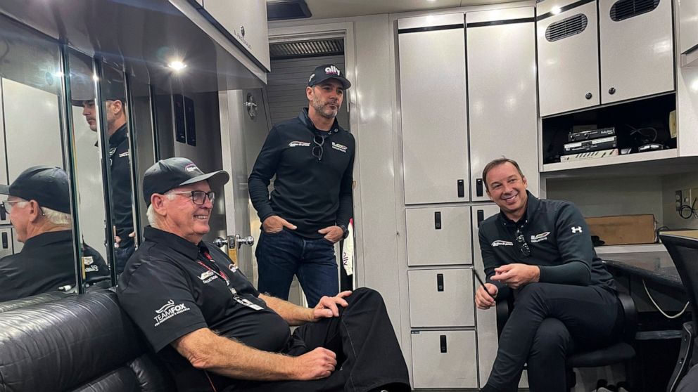 Seven-time NASCAR champion Jimmie Johnson stands in the doorway of the team hauler at Road Atlanta Raceway in Braselton, Georgia, Friday, Nov. 12, 2021, talking with Chad Knaus, right, his former crew chief, and Gary Nelson, team manager of the IMSA 