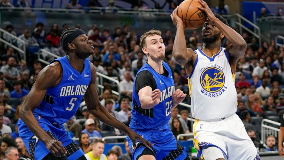 Golden State Warriors forward Andrew Wiggins, right, looks for a shot against Orlando Magic forward Franz Wagner, center, and center Mo Bamba (5) during the first half of an NBA basketball game, Tuesday, March 22, 2022, in Orlando, Fla. (AP Photo/Joh