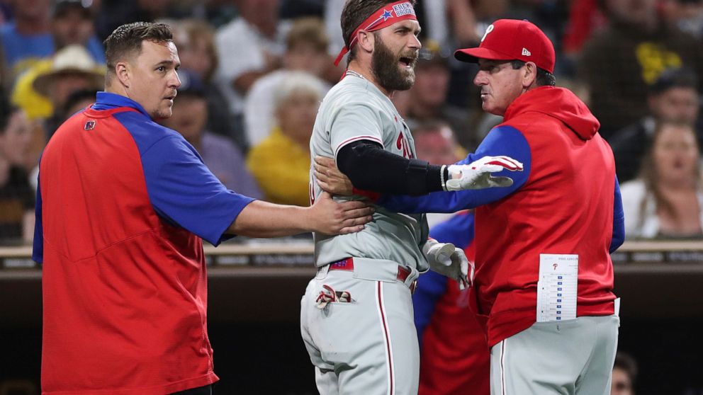 Philadelphia Phillies' Bryce Harper, center, reacts towards San Diego Padres pitcher Blake Snell after being hit by a pitch, as he walks off the field with interim manager Rob Thomson, right, and a trainer during the fourth inning of a baseball game 