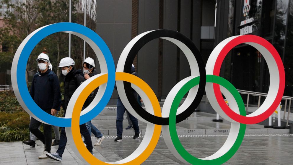 FILE - In this March 4, 2020, file photo, people wearing masks walk past the Olympic rings near the New National Stadium in Tokyo. It's been 2 1/2 months since the Tokyo Olympics were postponed until next year because of the COVID-19 pandemic. So whe
