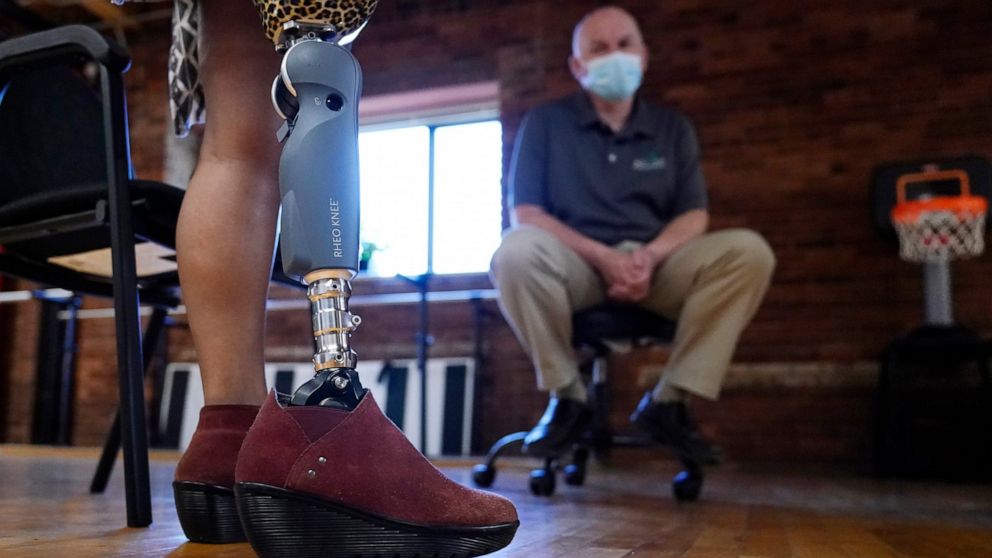 Claudine Humure, of Rwanda, takes a step with her new prosthetic leg as Arthur Graham, prosthetist at Next Step Bionics & Prosthetics, looks on, Monday, Sept. 20, 2021, in Newton, Mass. Humure was orphaned during her country's genocide and lost part 