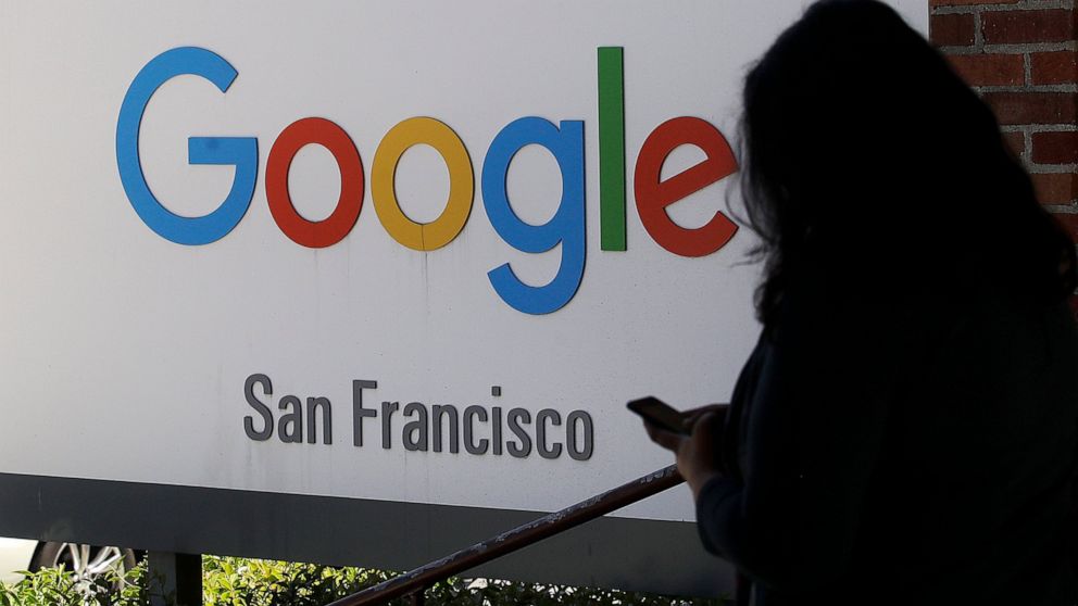 FILE - In this May 1, 2019, file photo, a woman walks past a Google sign in San Francisco. Google is making a $1 billion commitment to address the soaring price of housing in the San Francisco Bay Area, a problem that the internet company and its Sil