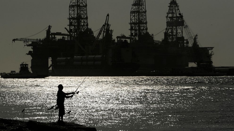FILE - A man wears a face mark as he fishes near docked oil drilling platforms, Friday, May 8, 2020, in Port Aransas, Texas. The U.S. Interior Department on Wednesday, Nov. 17, 2021, is auctioning vast oil reserves in the Gulf of Mexico estimated to 