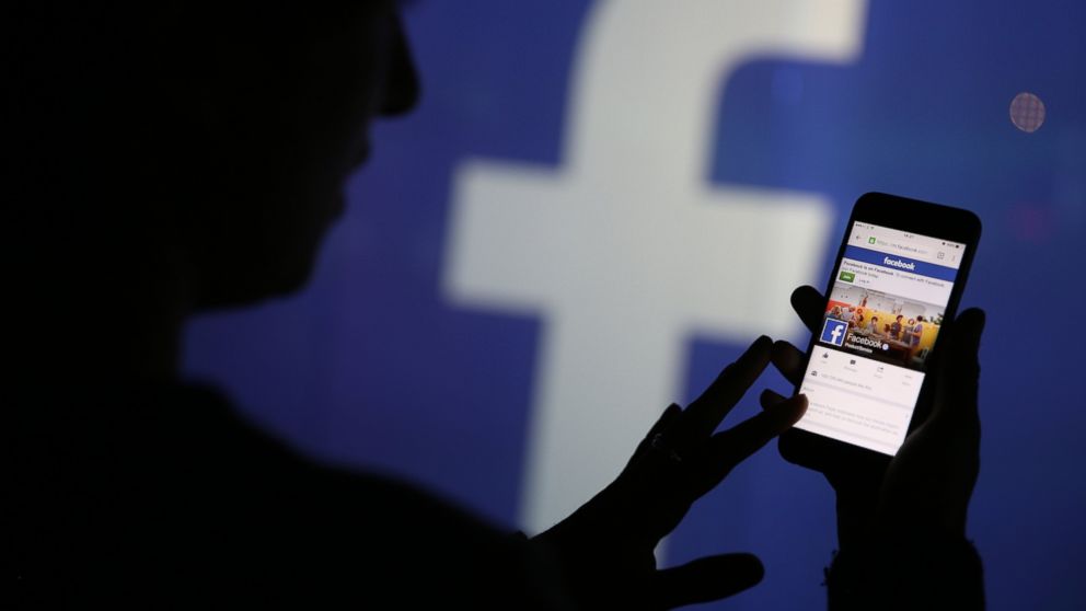 A woman checks the Facebook Inc. site on her smartphone while standing against an illuminated wall bearing the Facebook Inc. logo in this arranged photograph in London, Dec. 23, 2015.  