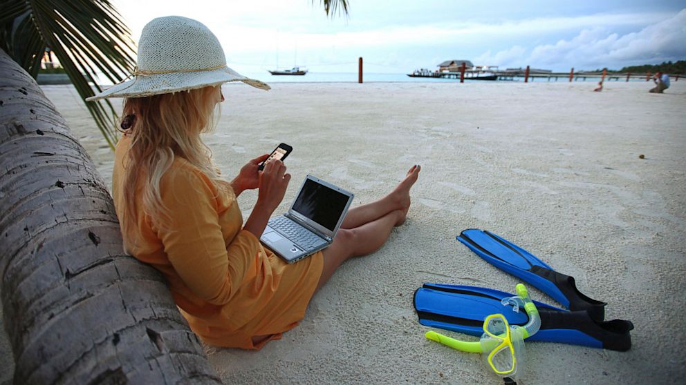 A woman sits on Villingili beach, working with a notebook and mobile phone.



